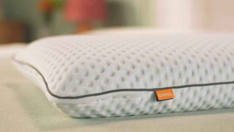 Oreiller blanc à surface texturée avec petits creux réguliers, bordé d’un passepoil gris et muni d’une étiquette orange “Emma” sur le côté, posé dans une chambre à la lumière douce.