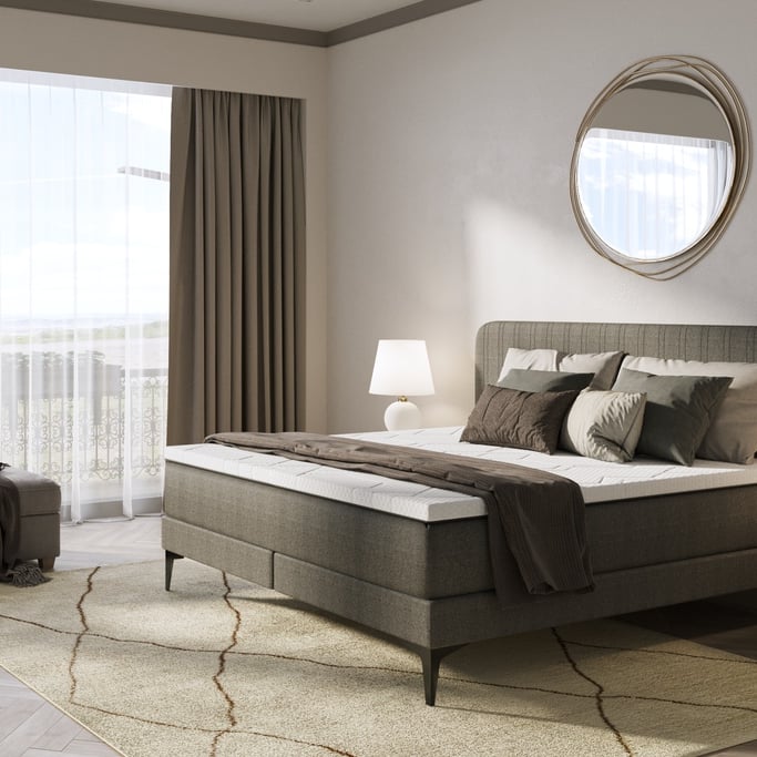 Modernes Schlafzimmer mit grauem Boxspringbett, Spiegel an der Wand und großen Fenstern mit Tageslicht.