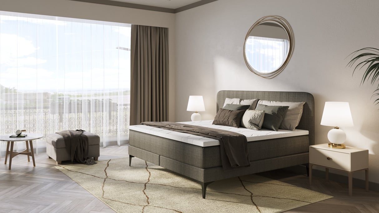 Modernes Schlafzimmer mit grauem Boxspringbett, Spiegel an der Wand und großen Fenstern mit Tageslicht.