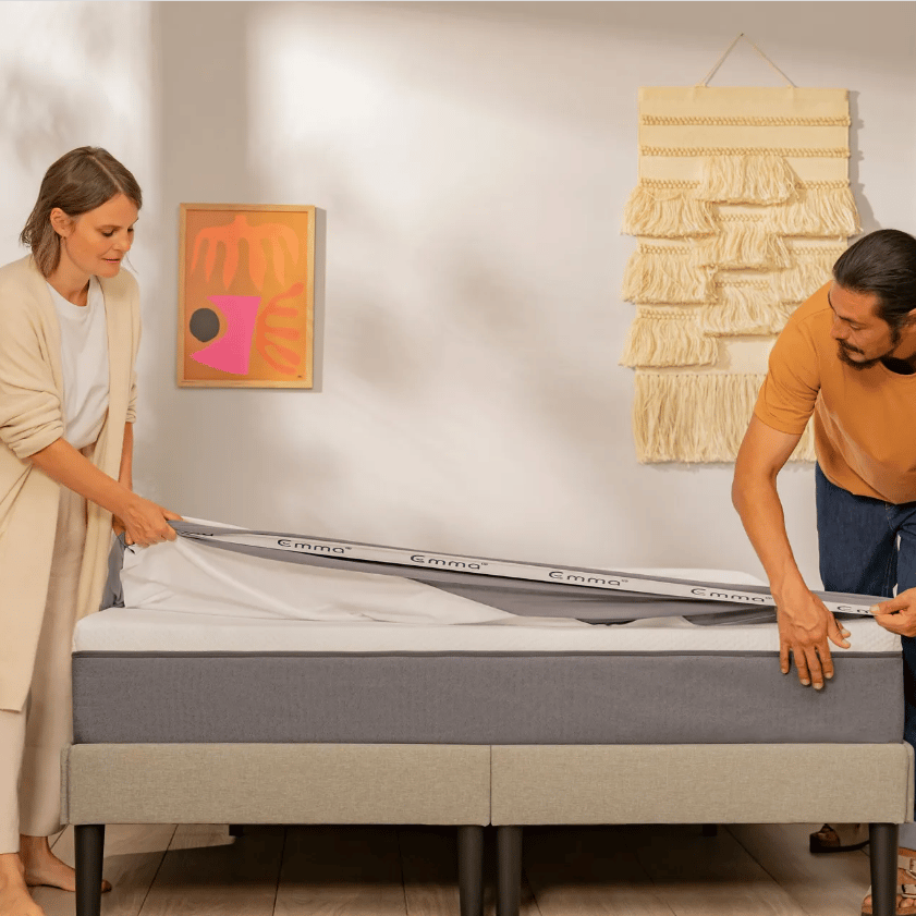 Twee personen plaatsen een matrasbeschermer van het merk Emma op een matras in een lichte kamer; links een groene kamerplant, rechts een beige wandhanger van geweven stof en abstract kunstwerk in roze- en oranjetinten; bedframe is grijs met zwarte poten, houten vloer zichtbaar.