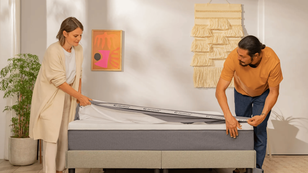 Twee personen plaatsen een matrasbeschermer van het merk Emma op een matras in een lichte kamer; links een groene kamerplant, rechts een beige wandhanger van geweven stof en abstract kunstwerk in roze- en oranjetinten; bedframe is grijs met zwarte poten, houten vloer zichtbaar.