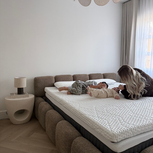 Twee jonge kinderen liggen op een royaal, lichtgekleurde matras terwijl een volwassene naast hen op bed leunt; de kamer is minimalistisch ingericht met een gestoffeerd bedframe, een klein nachttafeltje met lamp en zachte gordijnen waar daglicht doorheen valt.