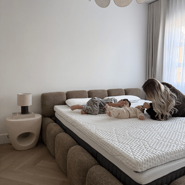 Twee jonge kinderen liggen op een royaal, lichtgekleurde matras terwijl een volwassene naast hen op bed leunt; de kamer is minimalistisch ingericht met een gestoffeerd bedframe, een klein nachttafeltje met lamp en zachte gordijnen waar daglicht doorheen valt.