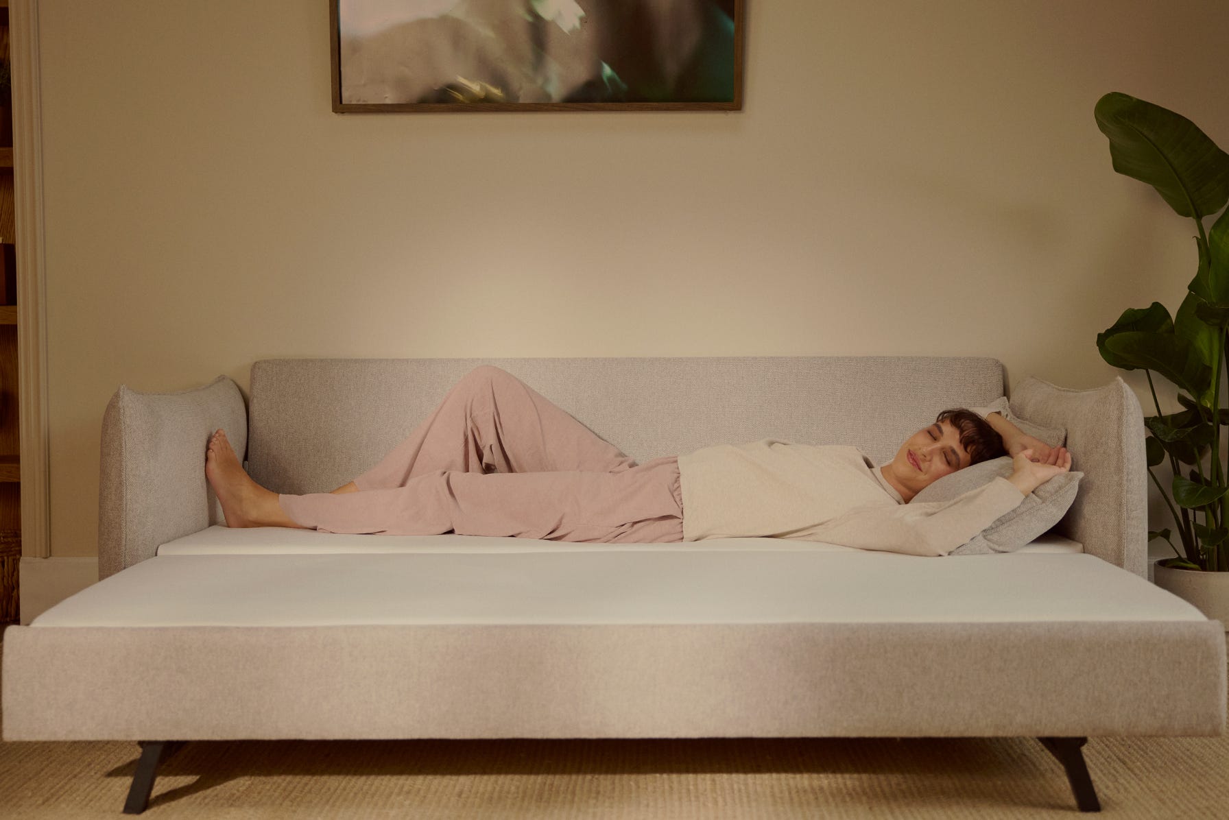 Canapé convertible gris avec deux coussins assortis, placé au centre d’un tapis clair dans un salon moderne. Devant, table basse en bois. À gauche, fauteuil marron à structure noire et grande plante verte en pot. Mur blanc avec deux affiches encadrées au-dessus du canapé. À droite, meuble bibliothèque intégré en bois avec livres, objets déco et plantes. Sol en bois chaud, lumière naturelle entrant par de grandes fenêtres avec rideaux blancs.