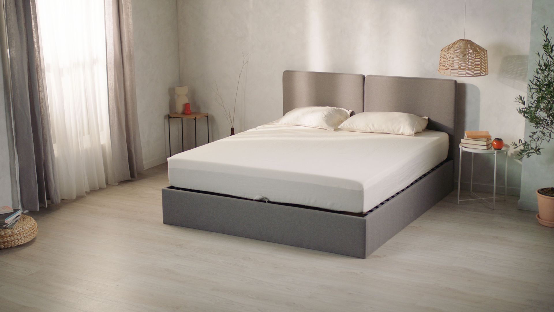 Lit Emma Coffre avec cadre et tête de lit rembourrés en gris, matelas blanc et deux oreillers crème. Placé dans une chambre lumineuse au sol en bois clair et murs gris doux. À gauche, rideaux blancs laissant filtrer la lumière naturelle. À droite, petite table ronde avec vase et livre. Suspension en rotin au-dessus du lit et plante verte dans le coin, créant une ambiance sereine et contemporaine.