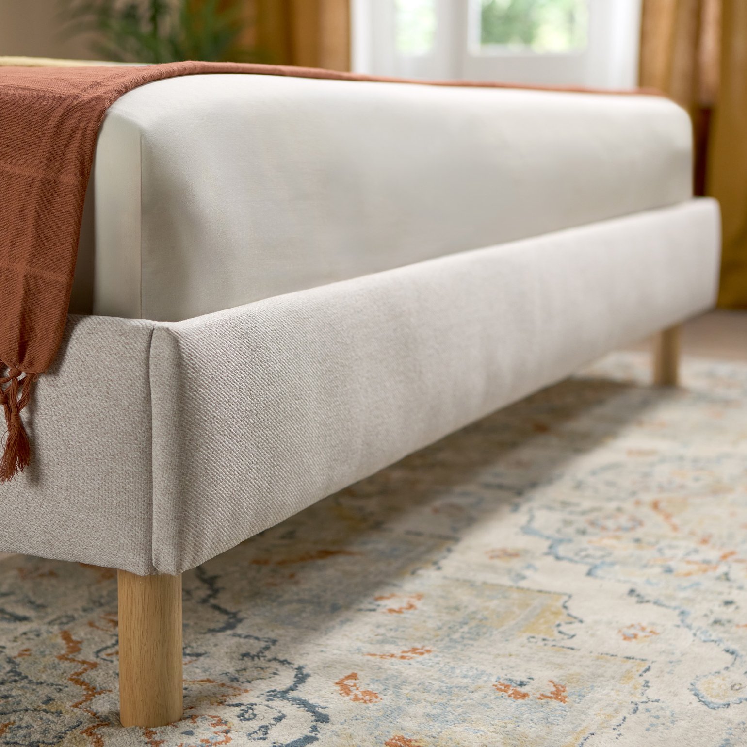 Emma bed in beige met gestoffeerd hoofdbord en houten poten, gestyled in een lichte, luchtige slaapkamer.