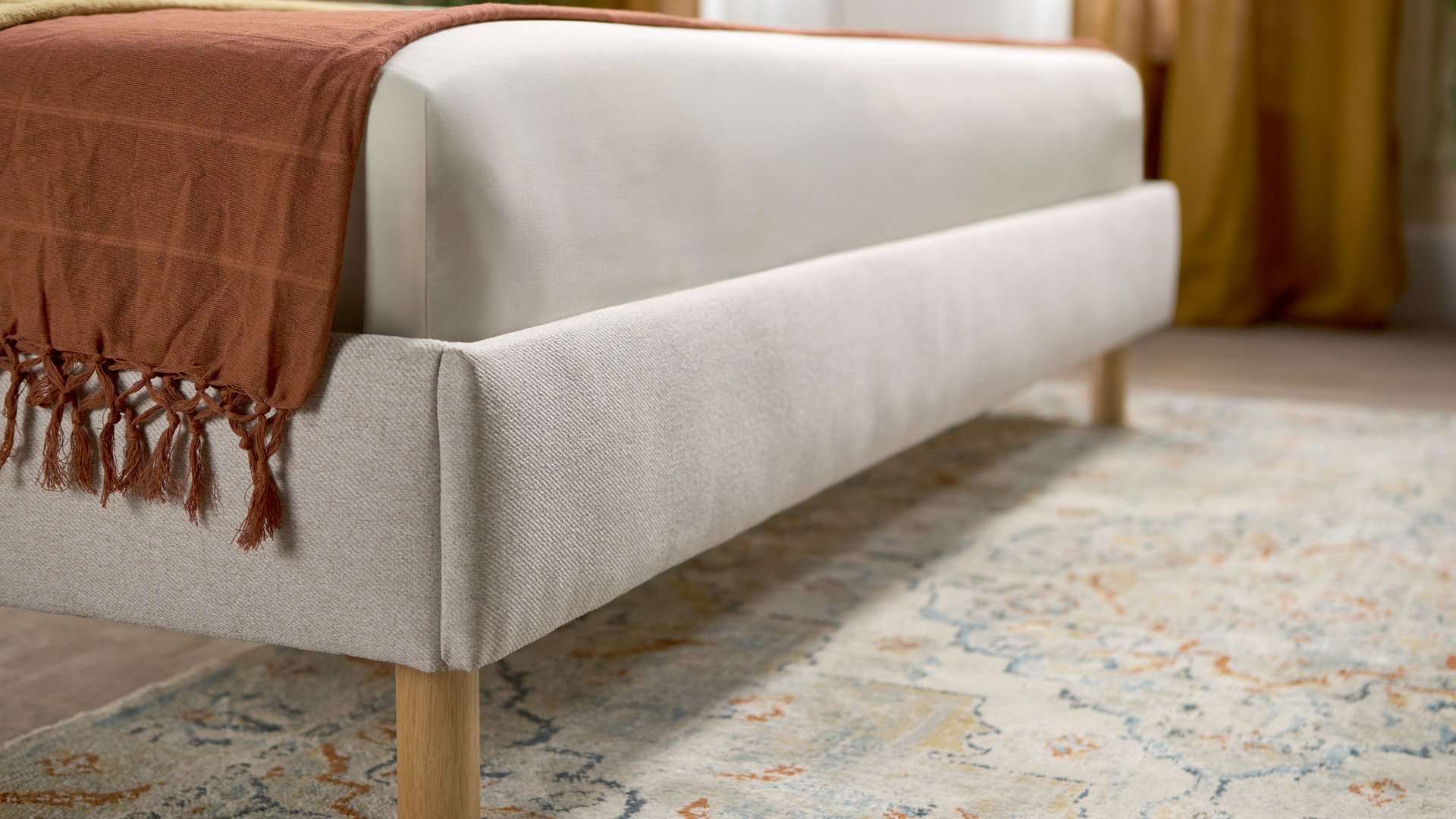 Emma bed in beige met gestoffeerd hoofdbord en houten poten, gestyled in een lichte, luchtige slaapkamer.