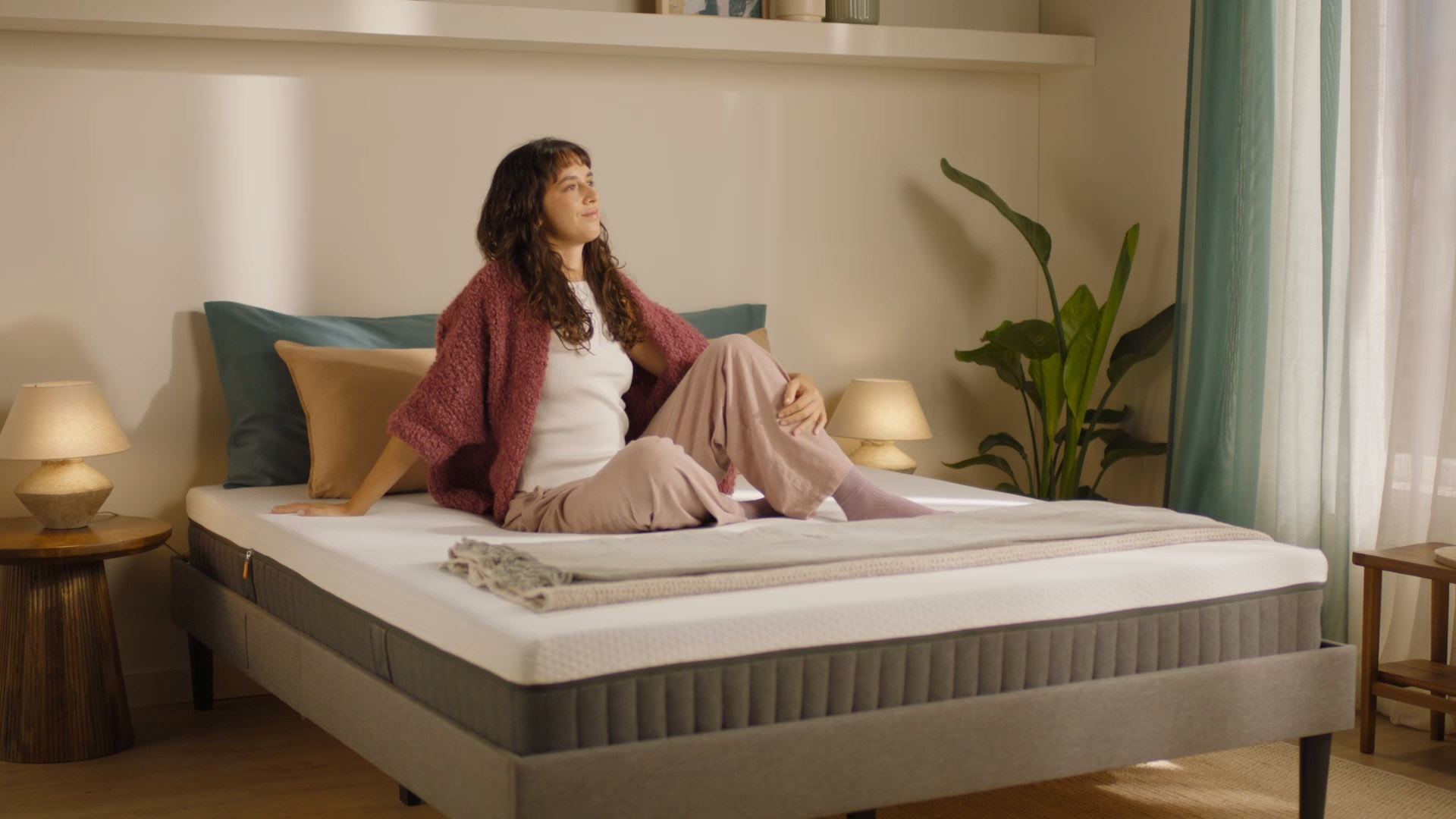 Person sitting on a grey upholstered bed with a hybrid mattress demonstrating everyday use.
