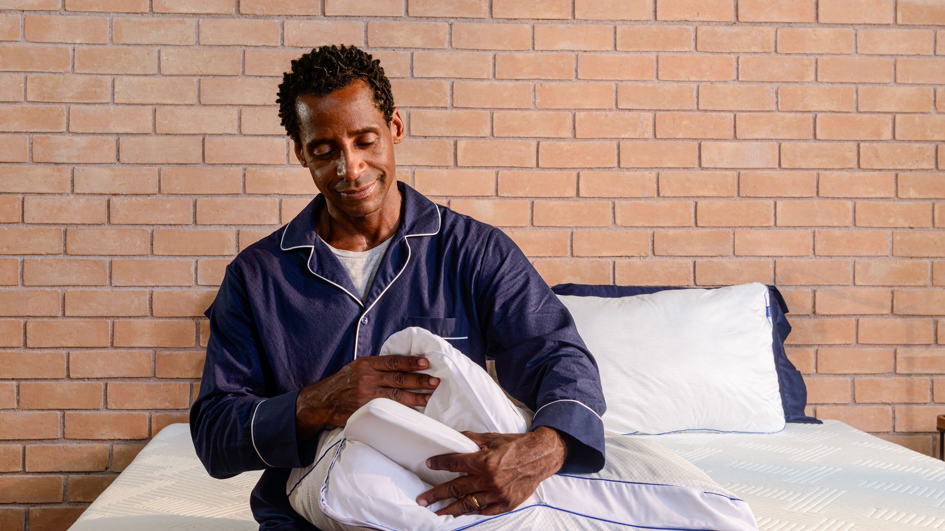 Homem sentado numa cama com fundo de parede de tijolos. Ele segura um travesseiro branco, abrindo-o para revelar as 3 camadas internas do travesseiro.