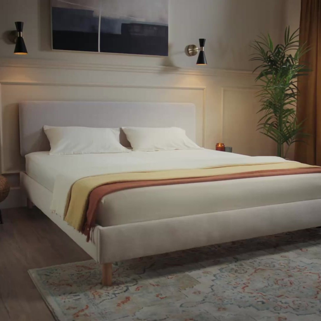 Emma Original Bed in light beige upholstered frame with natural wood legs in a dimly lit styled bedroom.