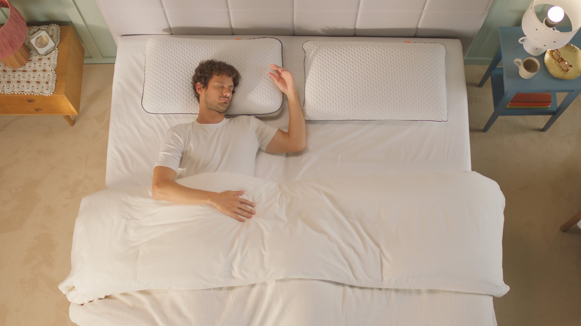 Persoon ligt onder een witte deken op een bed met twee witte Original Cooling Adapt Kussens met geribbeld oppervlak; links en rechts staan nachtkastjes met respectievelijk roze en witte lampen en decoratieve accessoires.