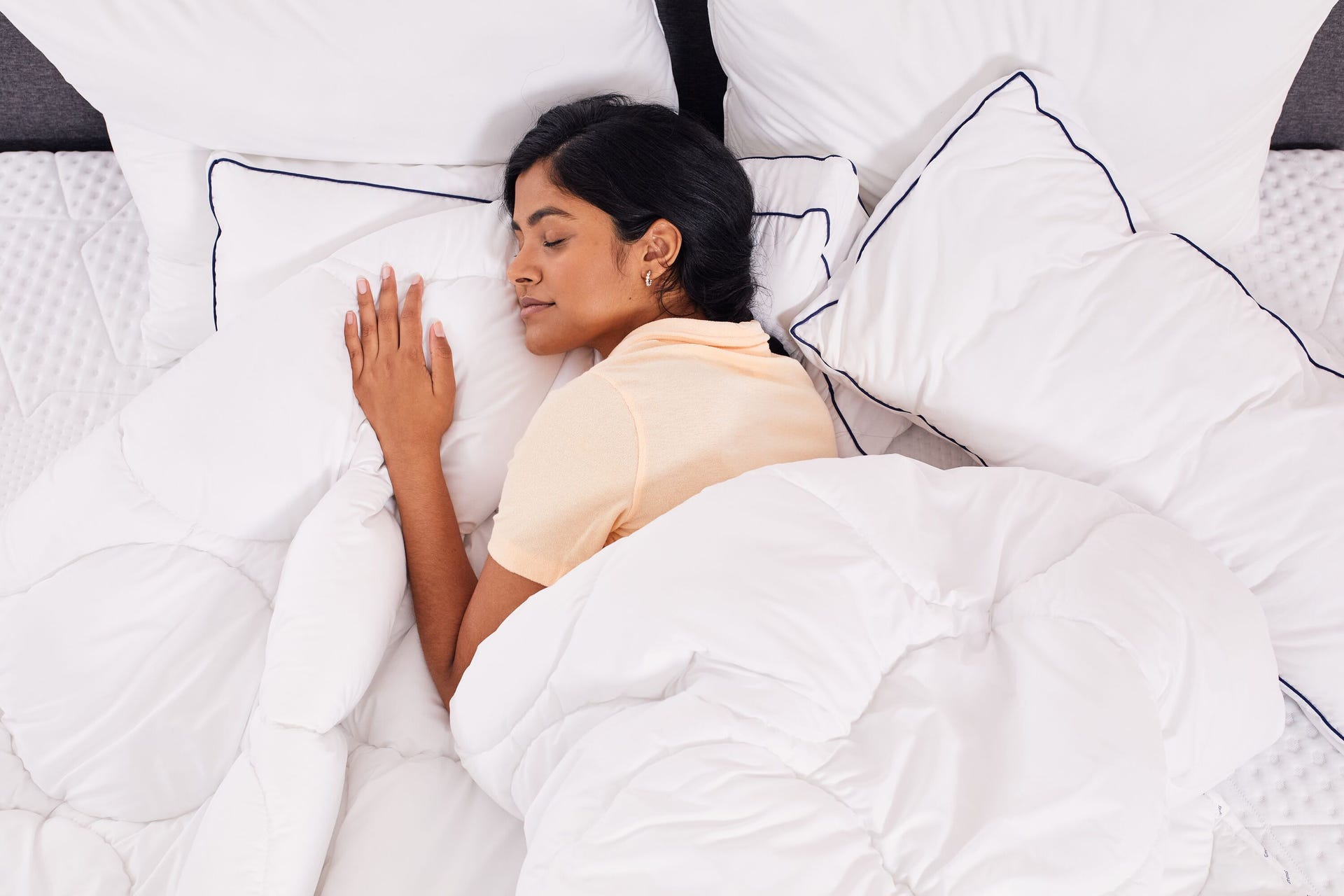Persoon liggend in bed onder een wit dekbed, gekleed in een licht shirt; hoofd rust op meerdere witte kussens met donkere biezen; matras is voorzien van een gewatteerde matrasbeschermer die links zichtbaar is; setting straalt comfort en rust uit.