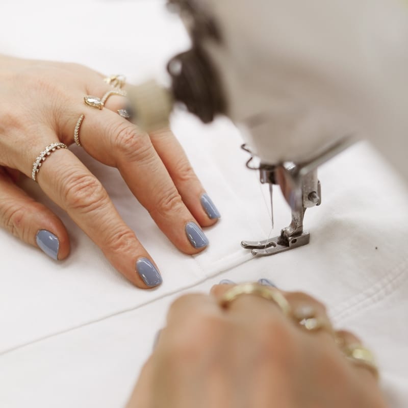 Close-up van handen met blauwe nagellak die wit textiel onder een naald van een naaimachine begeleiden—focus op precisie en ambacht in textielbewerking.