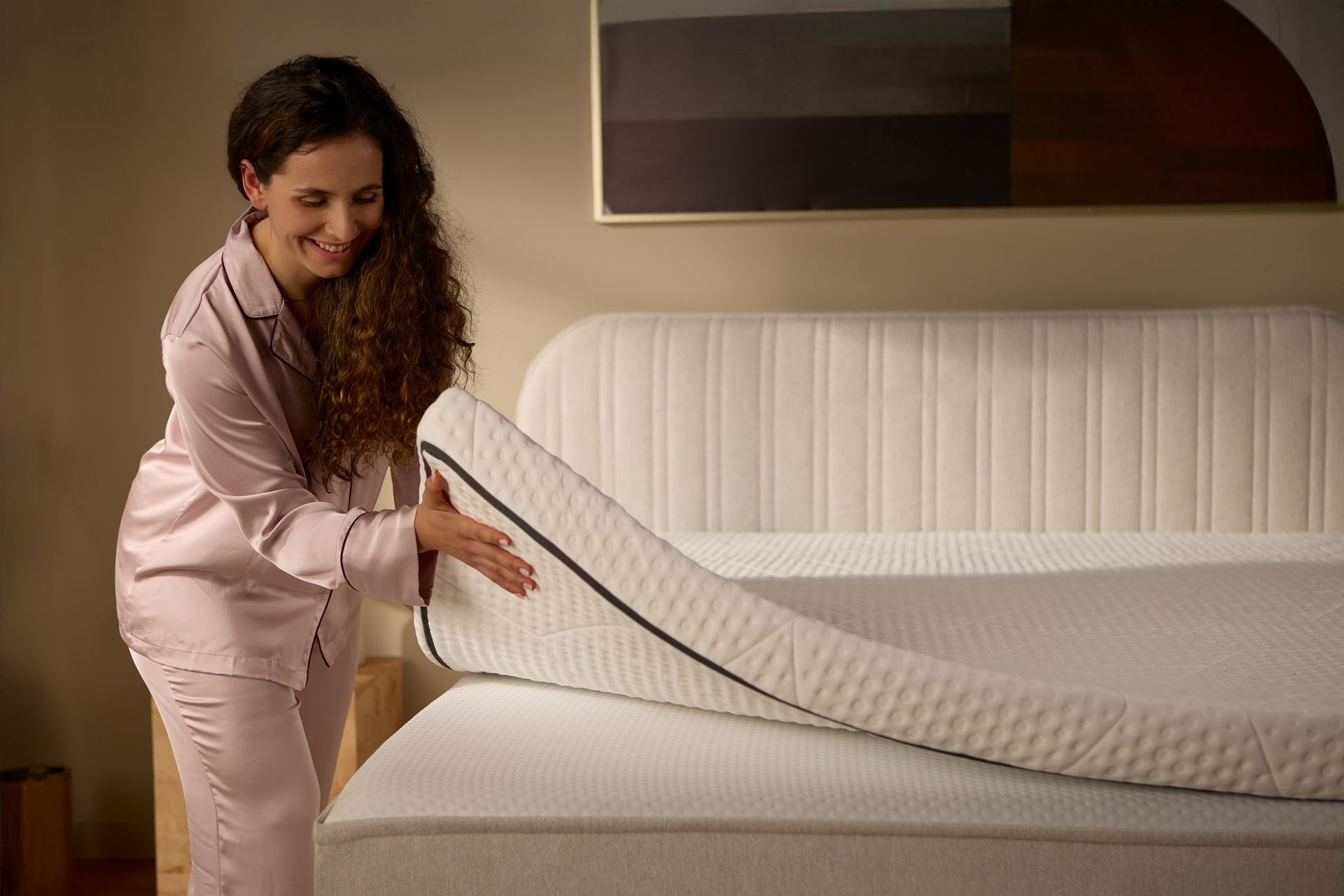Detail view with a person lifting a separate mattress topper placed on the bed, showing its thickness and flexible construction.