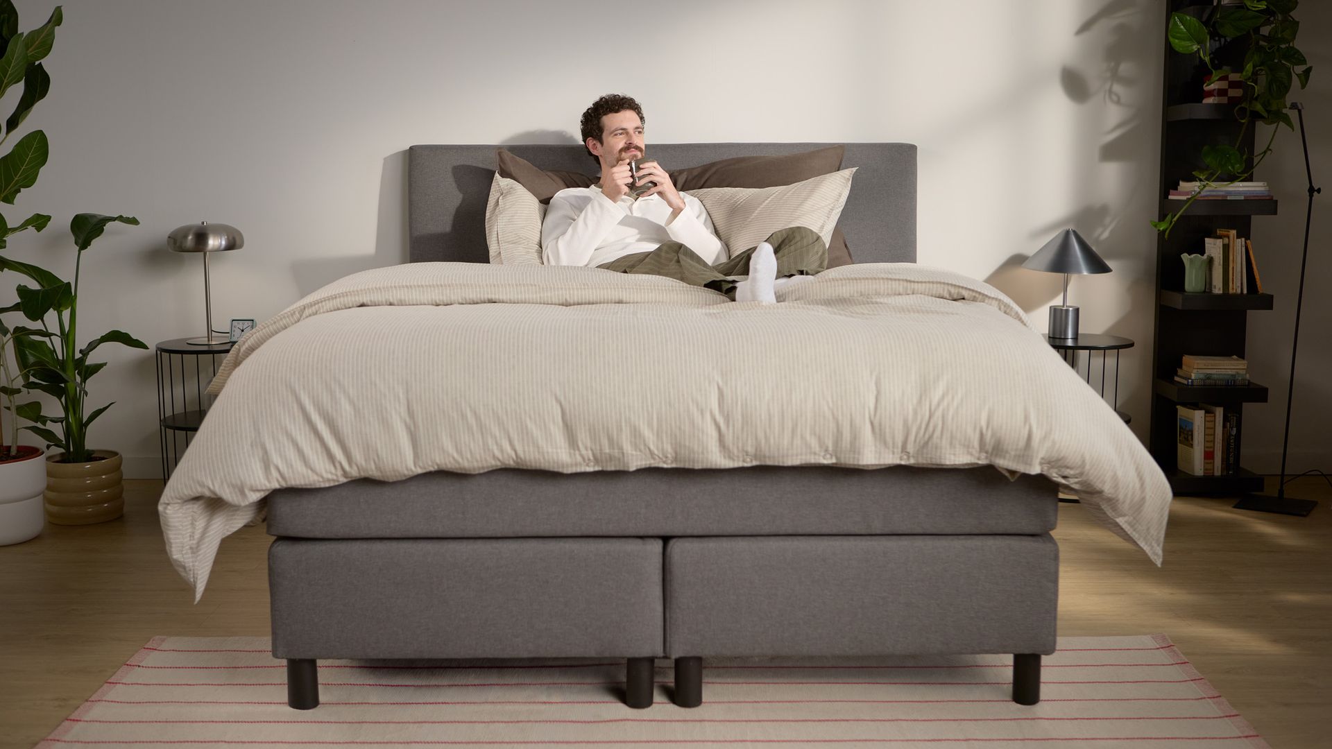Lifestyle beeld met een persoon die ontspant; toont gelaagd beddengoed en ondersteuning van het hoofdbord, met meerdere zichtbare steunpoten in het midden en langs de randen.