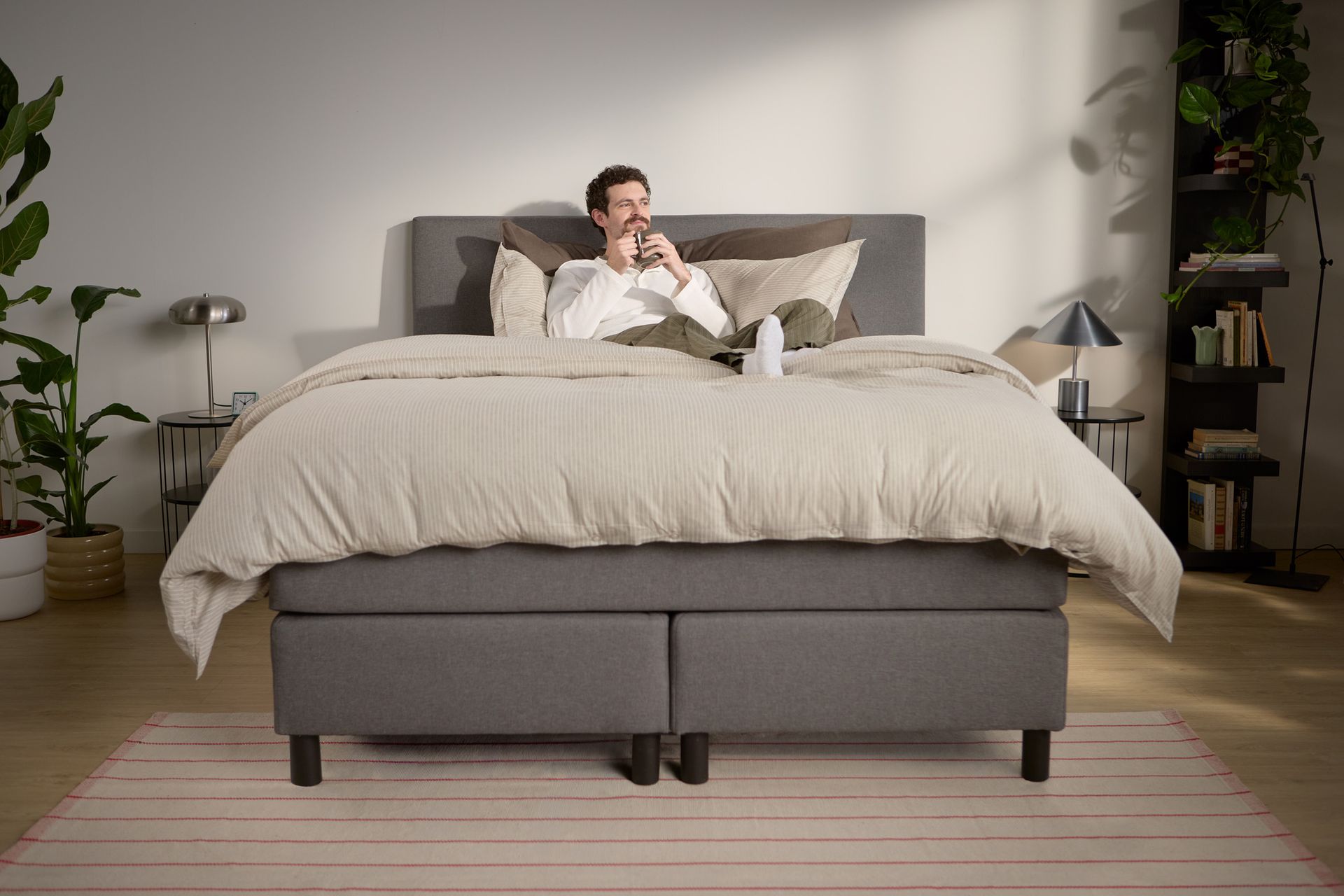 Lifestyle beeld met een persoon die ontspant; toont gelaagd beddengoed en ondersteuning van het hoofdbord, met meerdere zichtbare steunpoten in het midden en langs de randen.