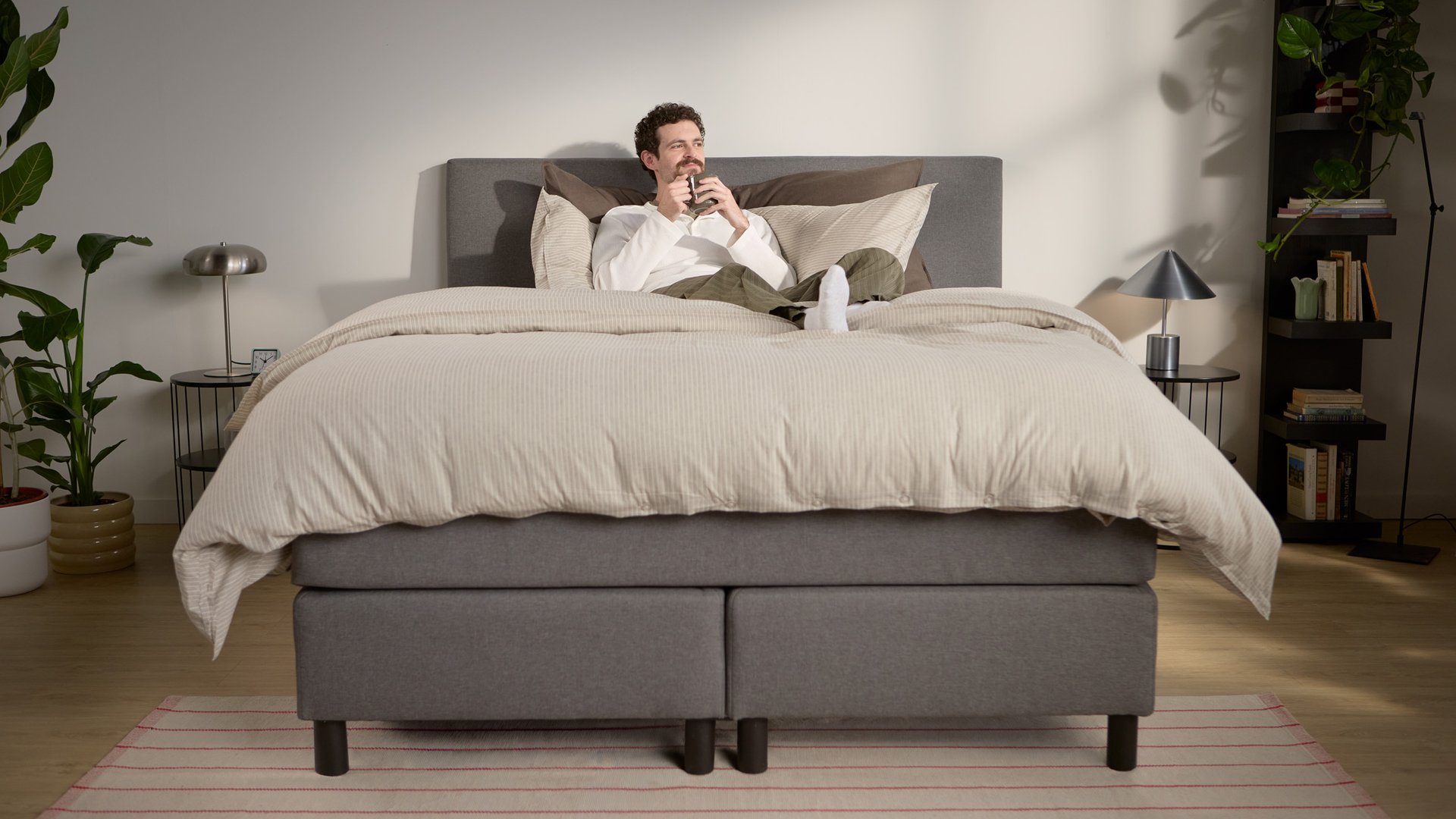 Lifestyle-Ansicht mit einer entspannten Person; zeigt mehrlagige Bettwäsche und die Unterstützung des Kopfteils, mit mehreren sichtbaren Stützbeinen in der Mitte und an den Rändern.