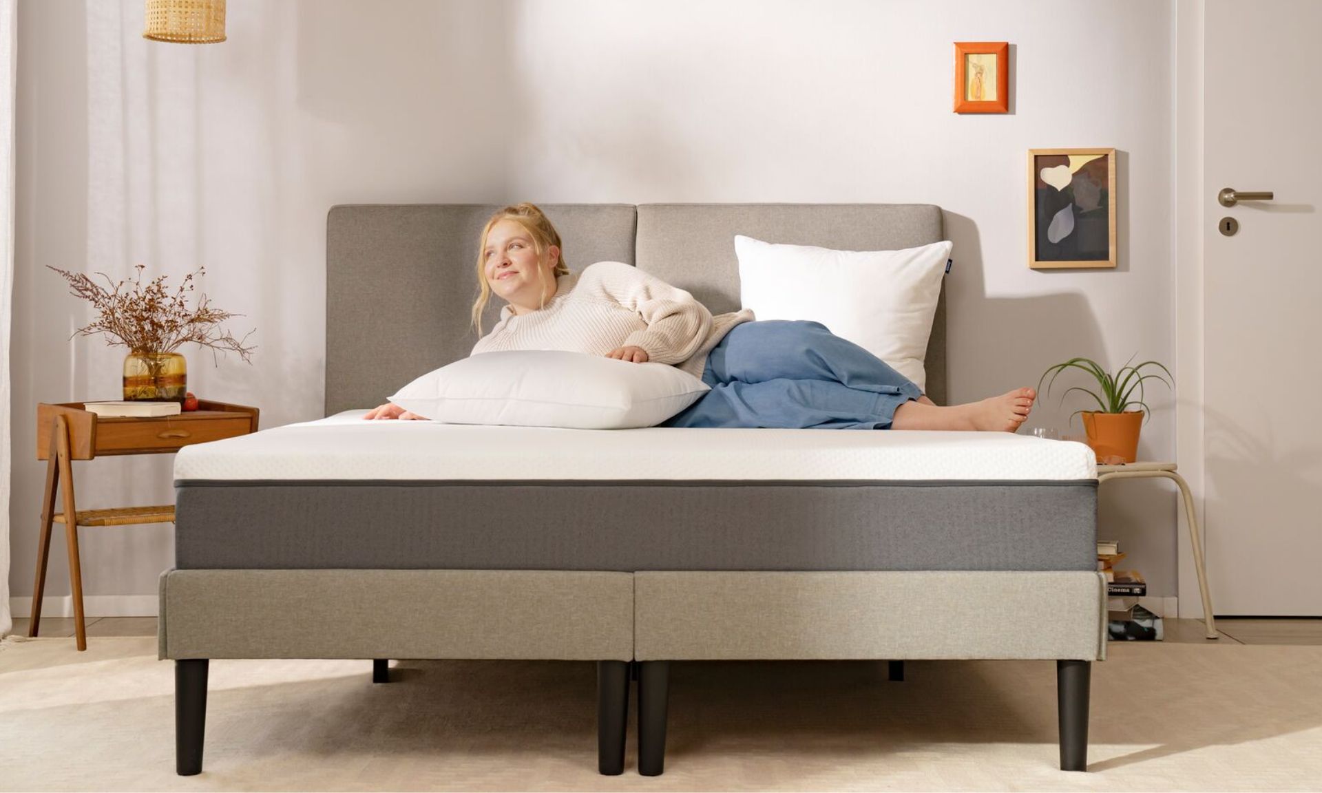 Persona sdraiata su letto con testiera grigia, cuscini bianchi e grigi, comodini con pianta e fiori secchi, in camera minimalista