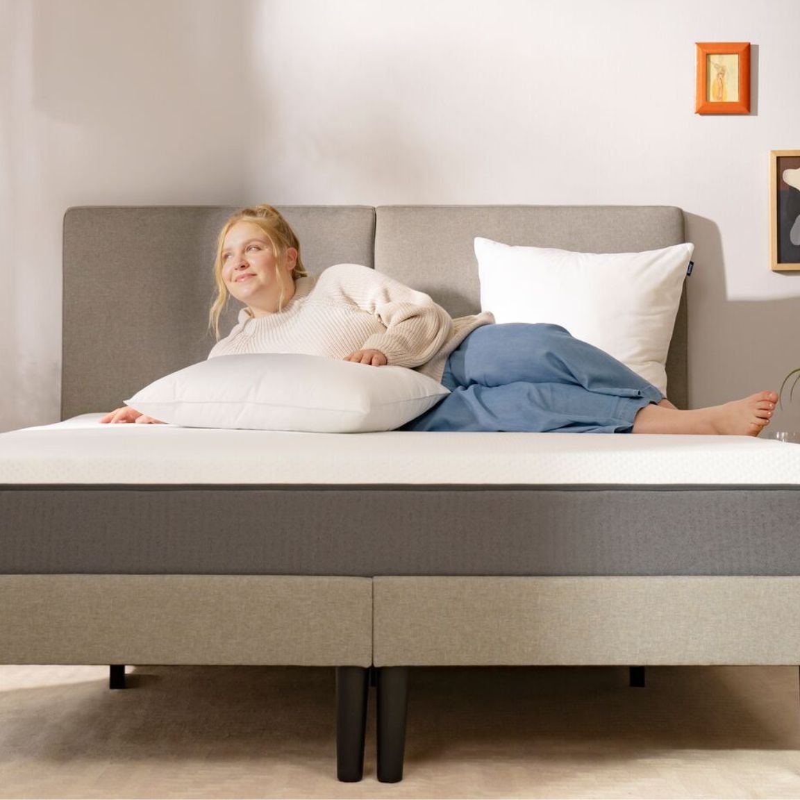 Persona sdraiata su letto con testiera grigia, cuscini bianchi e grigi, comodini con pianta e fiori secchi, in camera minimalista