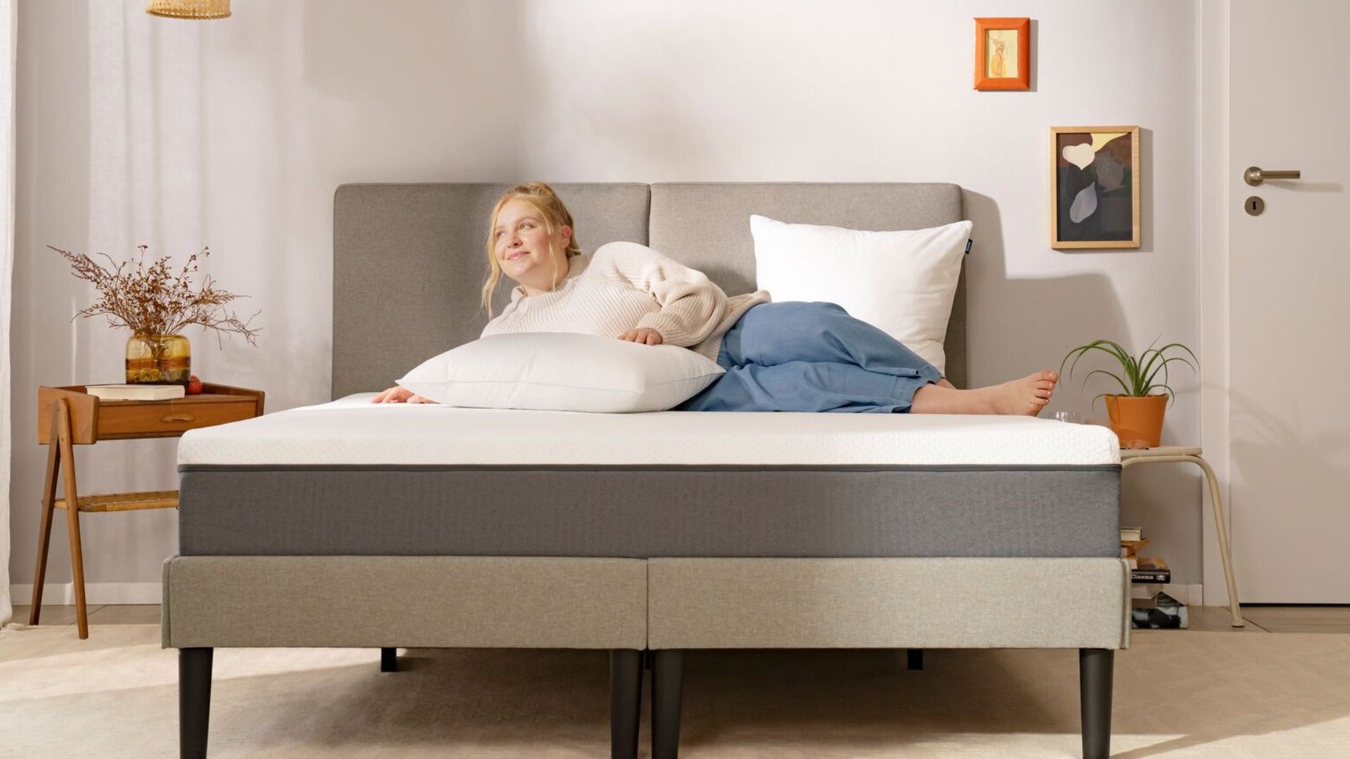 Persona sdraiata su letto con testiera grigia, cuscini bianchi e grigi, comodini con pianta e fiori secchi, in camera minimalista