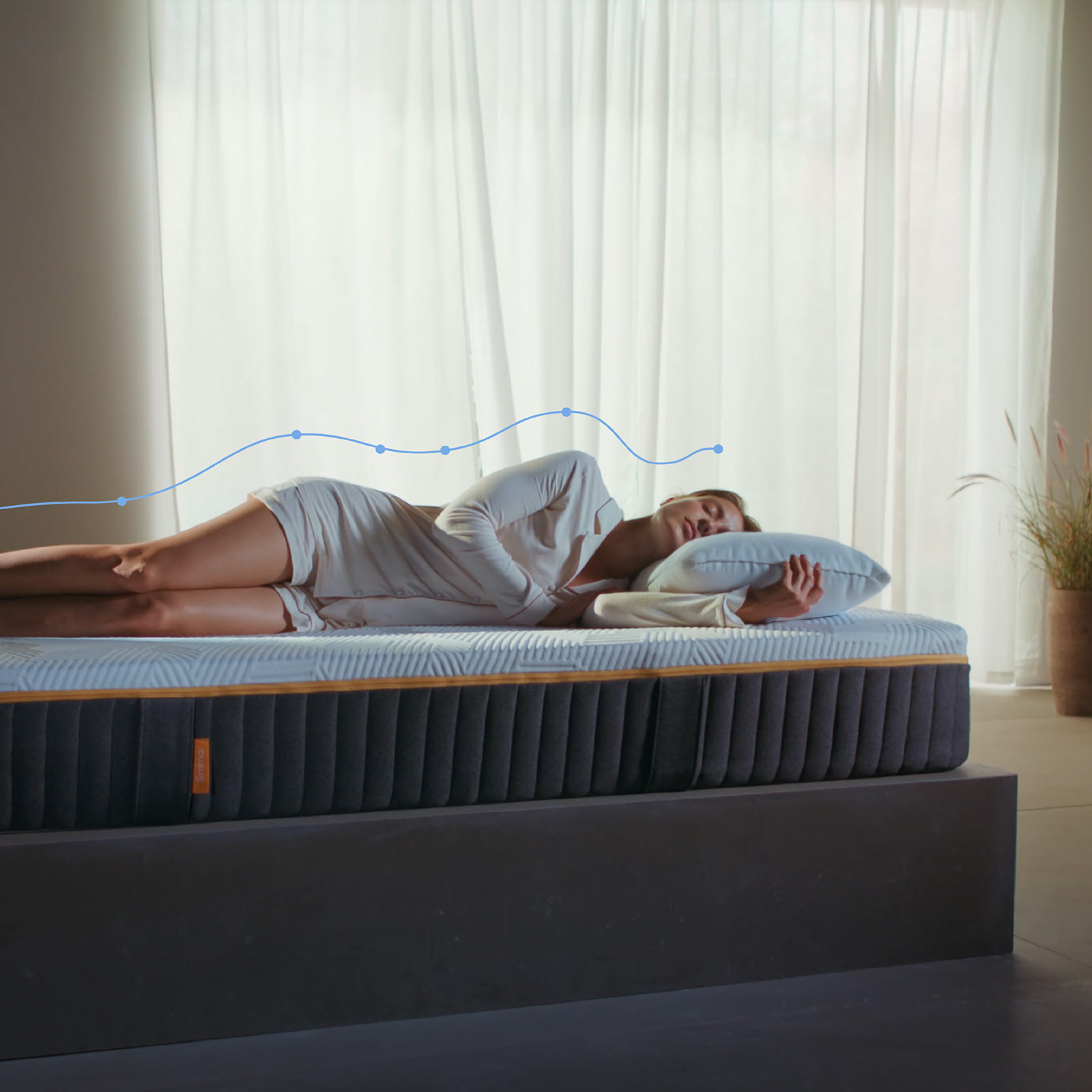 Mulher deitada de lado num colchão Emma Original Elite, com uma sobreposição gráfica de alinhamento da coluna a mostrar o suporte do corpo, num quarto luminoso.
