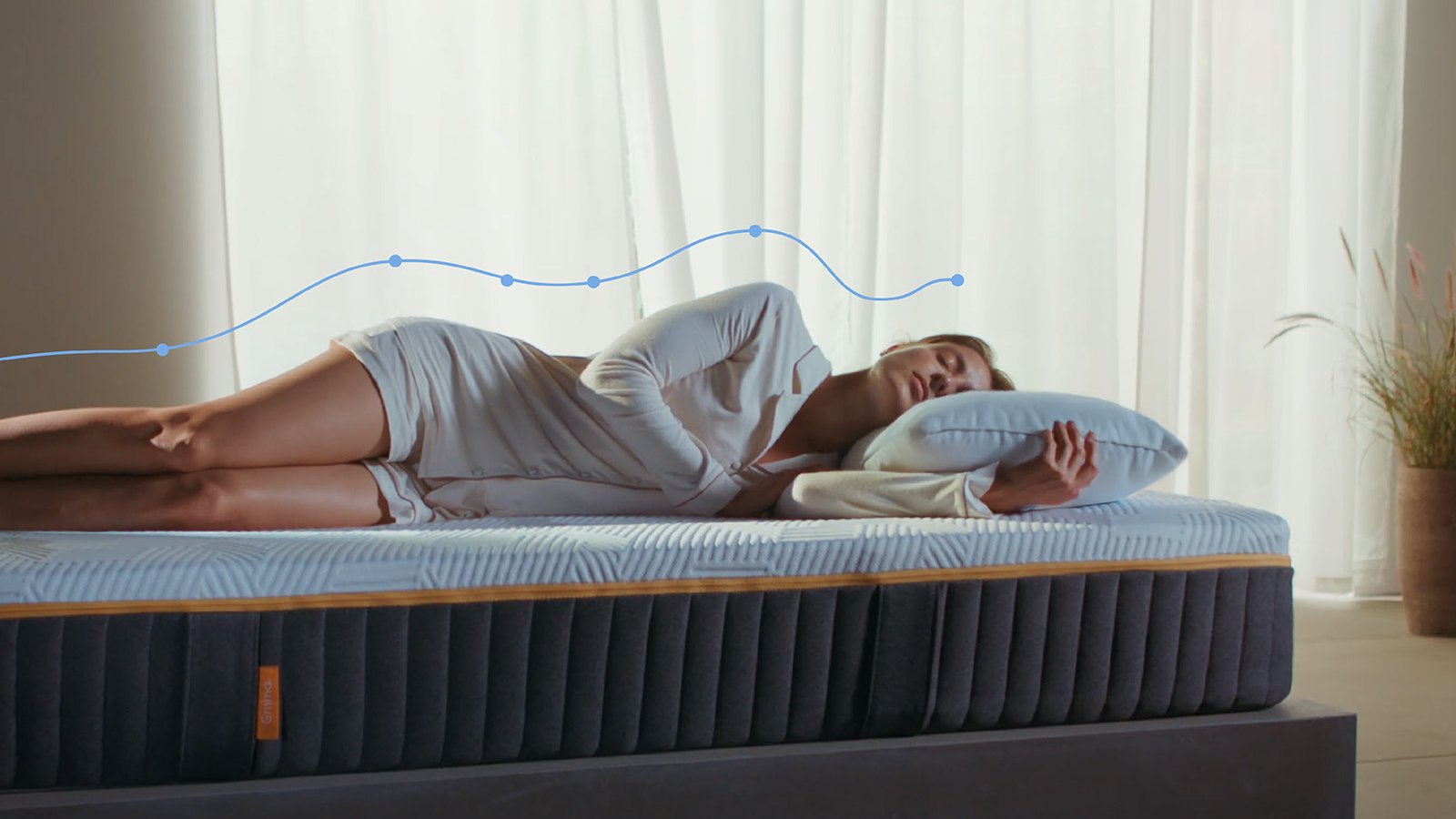 Mulher deitada de lado num colchão Emma Original Elite, com uma sobreposição gráfica de alinhamento da coluna a mostrar o suporte do corpo, num quarto luminoso.