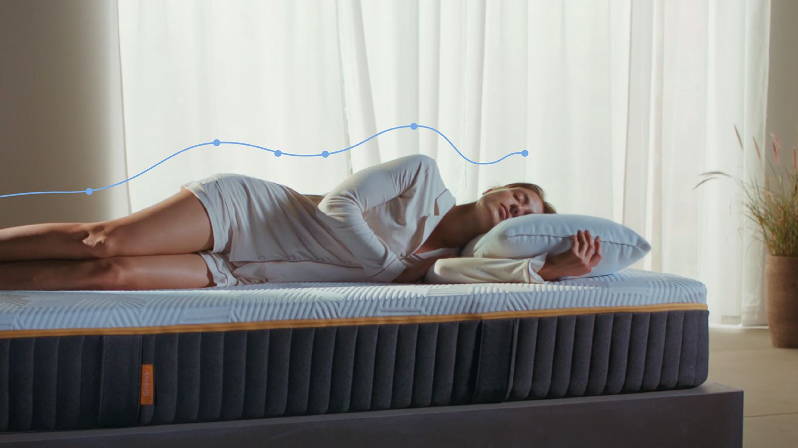 Mulher deitada de lado num colchão Emma Original Elite, com uma sobreposição gráfica de alinhamento da coluna a mostrar o suporte do corpo, num quarto luminoso.