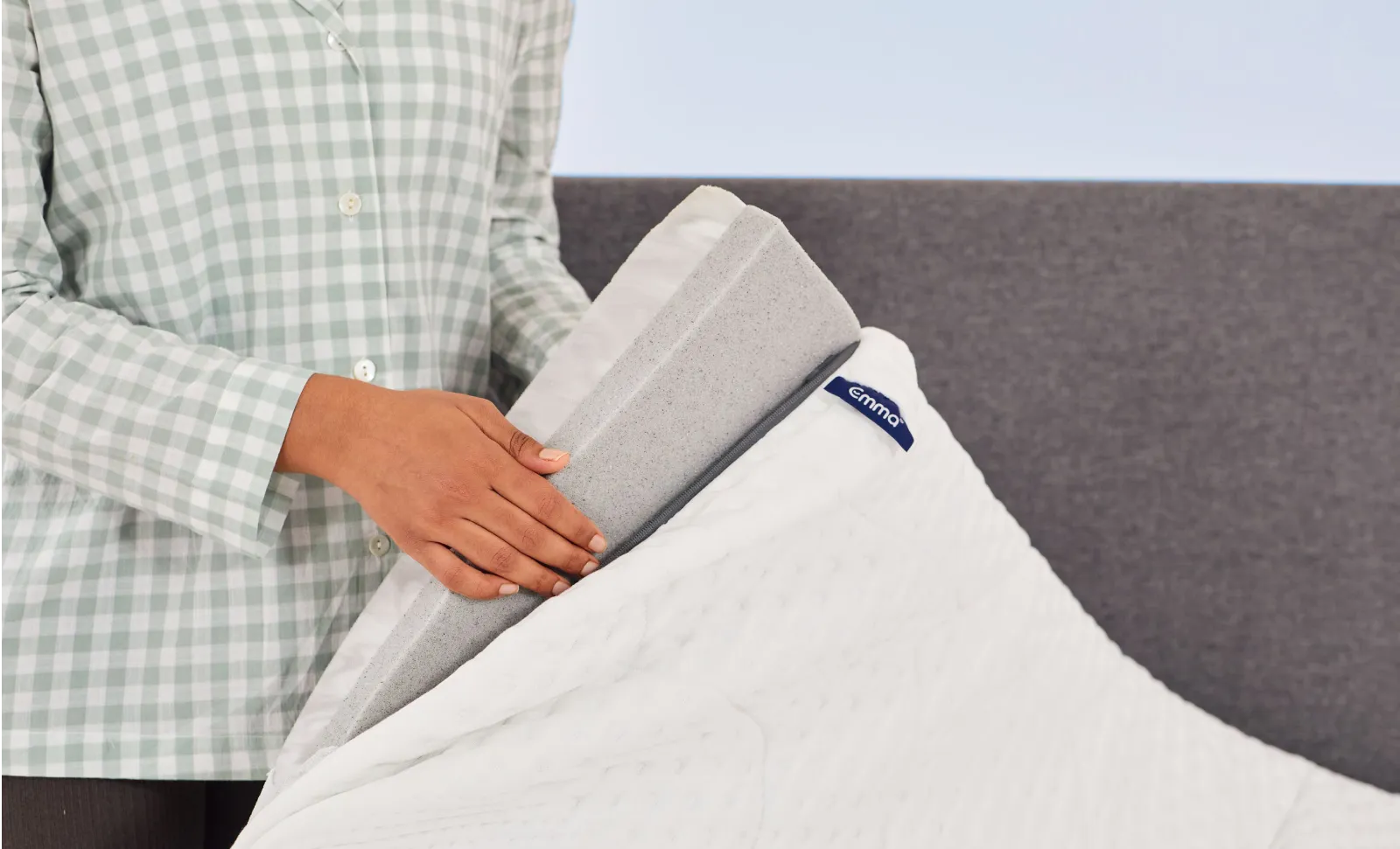 Personne portant un t-shirt vert ouvrant un surmatelas, montrant ses couches et le logo bleu Emma, sur un fond gris et blanc.