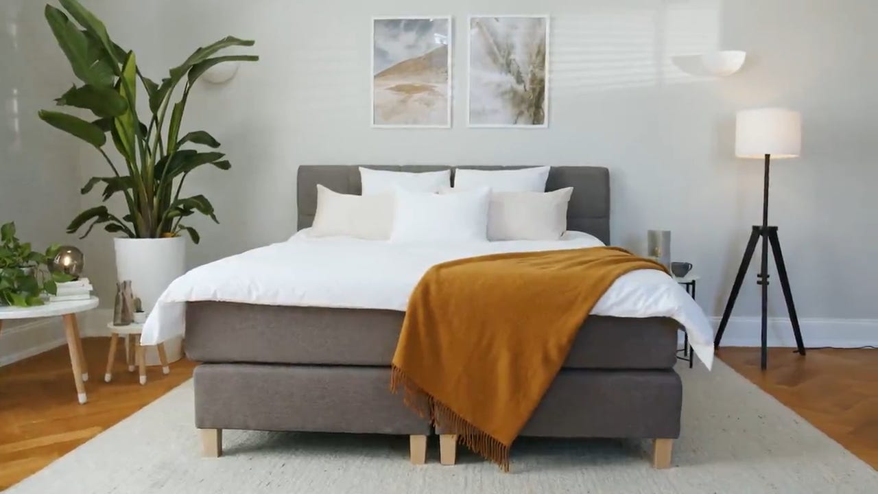 Emma Deluxe Boxspring in moderne slaapkamer met donkergrijs gestoffeerd frame en wit matras; bed op houten poten, opgemaakt met witte lakens en beige en witte kussens, mosterdgele plaid aan voeteneind; decoratieve elementen zoals planten, wandkunst, lampen en bijzettafels benadrukken stijlvolle en warme sfeer.