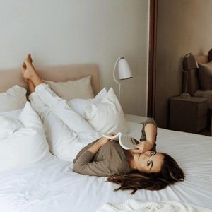 Mulher sorridente deitada de lado sobre cama com travesseiro Emma em quarto iluminado