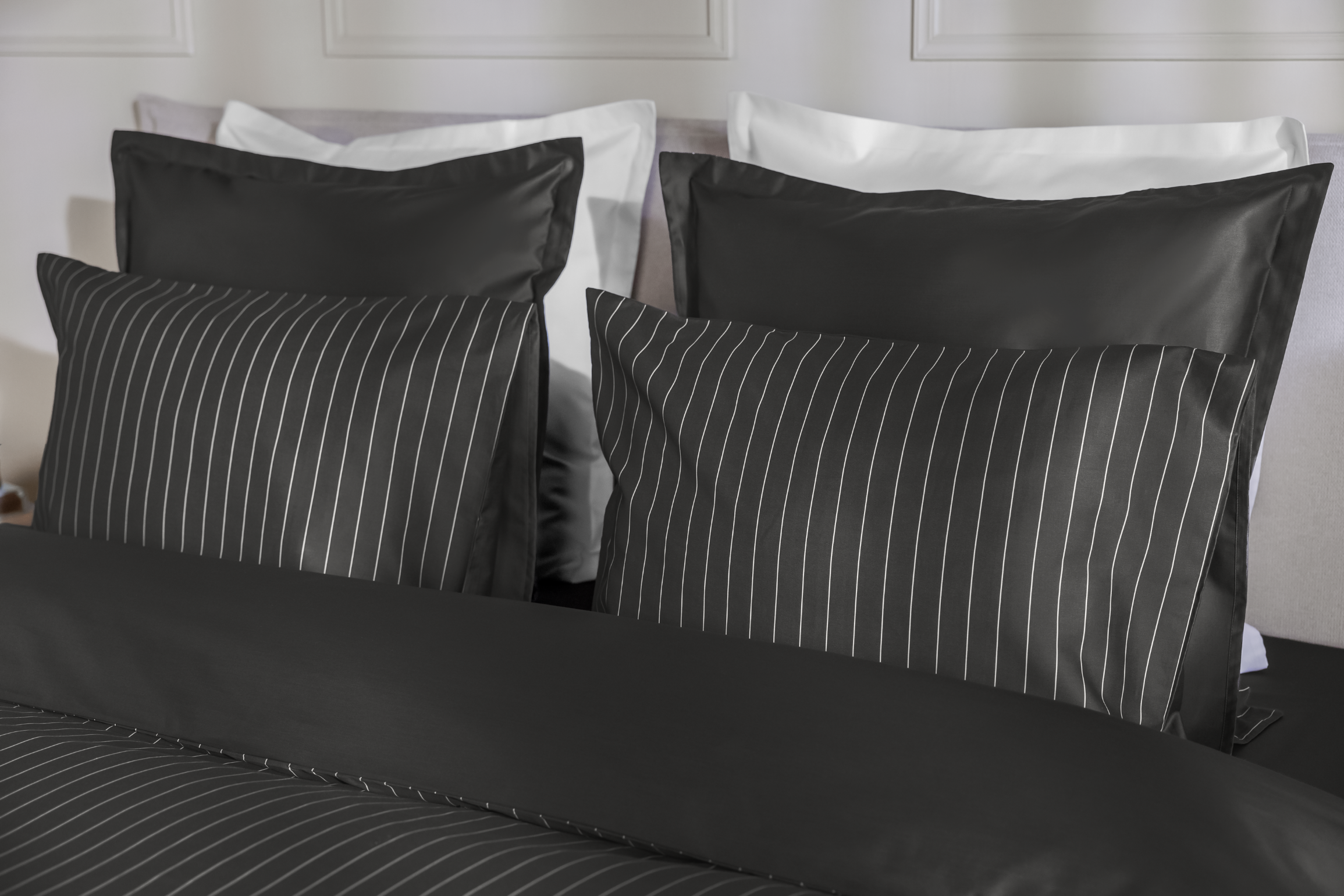 Detail van een bedhoek met een Emma Satijn Hoeslaken in grijs met subtiele verticale witte strepen, strak om een matras gespannen; hoeslaken heeft een lichte glans en gladde afwerking die het gestreepte patroon elegant accentueert; bedframe is grijs met houten poten; de setting is modern en minimalistisch met een witte muur, een lamp met gouden voet links, en droogbloemen aan de rechterzijde.