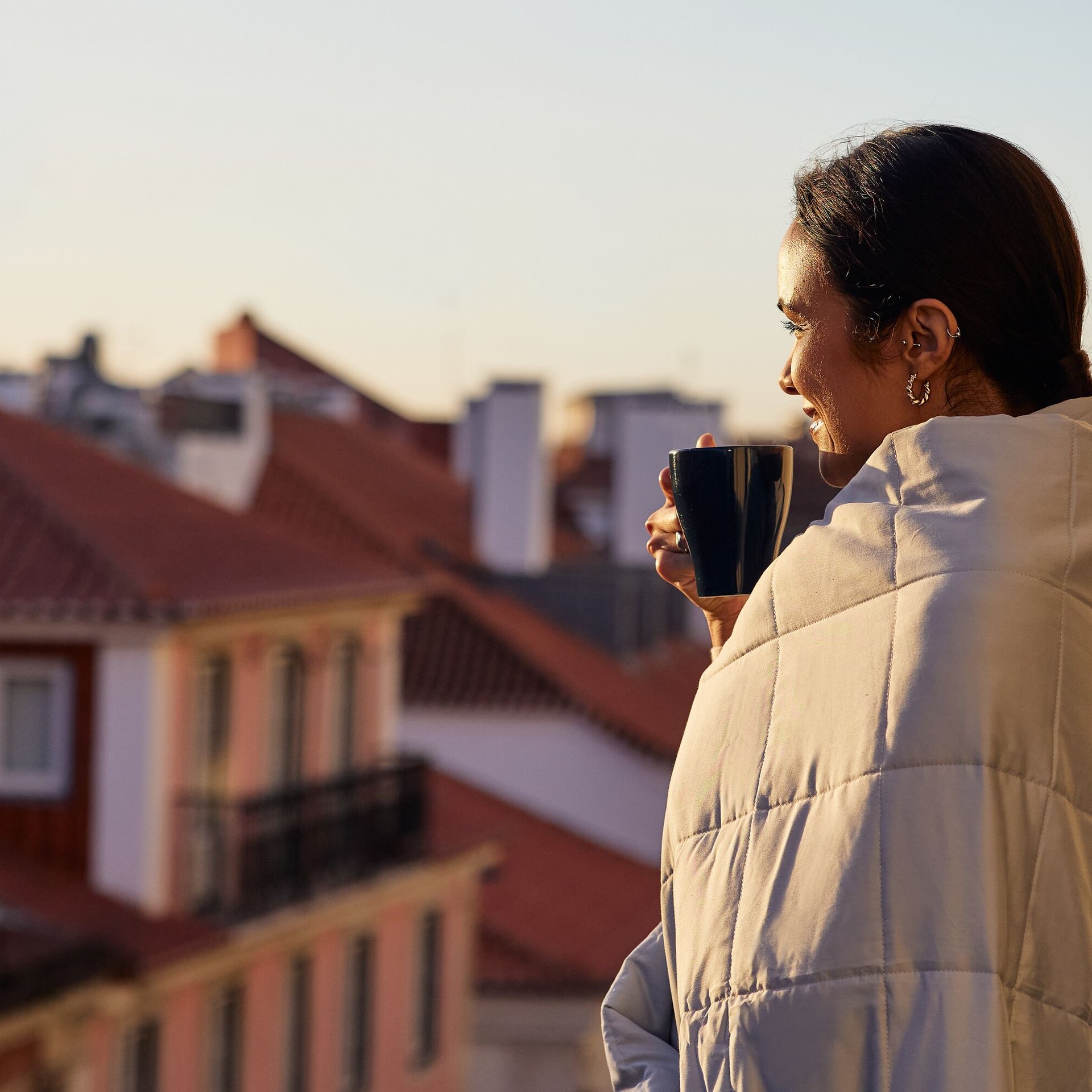 Persoon staat op een balkon gewikkeld in een lichtgekleurde Emma Verzwaringsdeken, met een zwarte mok in de hand, uitkijkend over een stad met rode daken bij zonsopgang – straalt rust en geborgenheid uit.