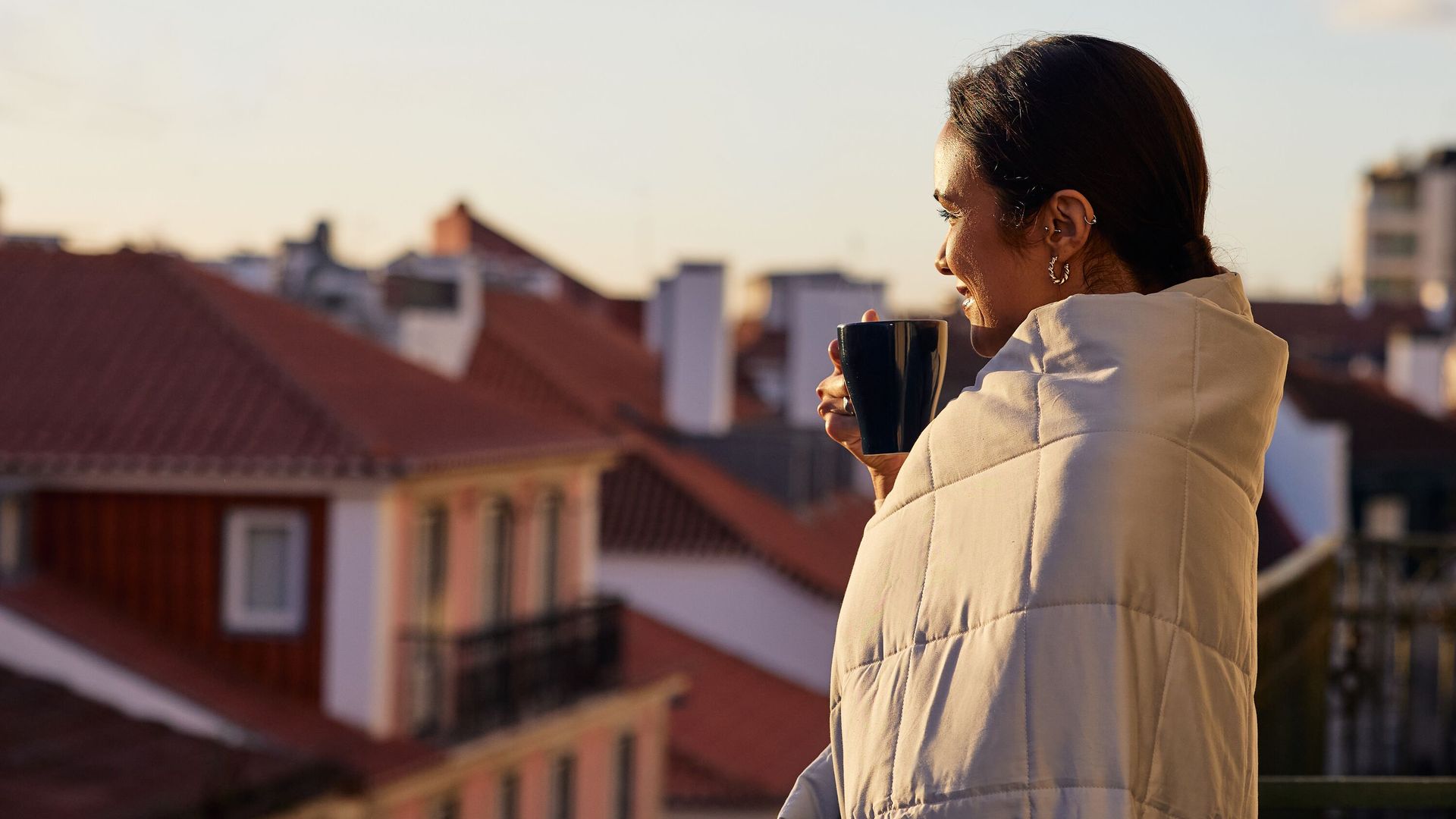 Persoon staat op een balkon gewikkeld in een lichtgekleurde Emma Verzwaringsdeken, met een zwarte mok in de hand, uitkijkend over een stad met rode daken bij zonsopgang – straalt rust en geborgenheid uit.