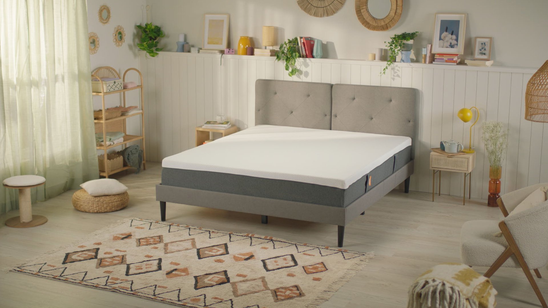 Camera accogliente con letto grigio, tappeto decorativo, tavolini con piante e libri, e luce naturale da tende bianche