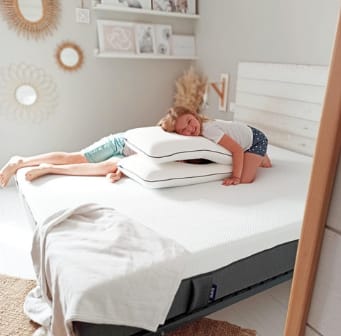 Twee kinderen op Emma matras spelen met Emma Diamond kussens in een lichte slaapkamer met houten vloer, beige tapijt en decoratieve wandspiegels.