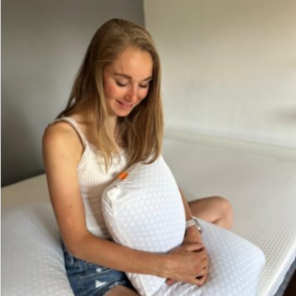 Vrouw zit ontspannen op bed en houdt het Emma AirGrid Wolkenkussen vast - een ergonomisch wit kussen met blauwe bies, ontworpen voor ademend comfort.