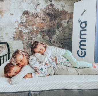 Three children lying on a mattress in a playful stack, with an "Emma" branded box beside the bed and a world map mural on the wall behind them.