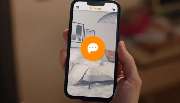Hand houdt smartphone vast met Emma-website geopend. Op het scherm is een slaapkamer te zien met opgemaakt bed, wanddecoratie en lamp. Een chaticoon en winkelwagenknop benadrukken interactie en online winkelen.