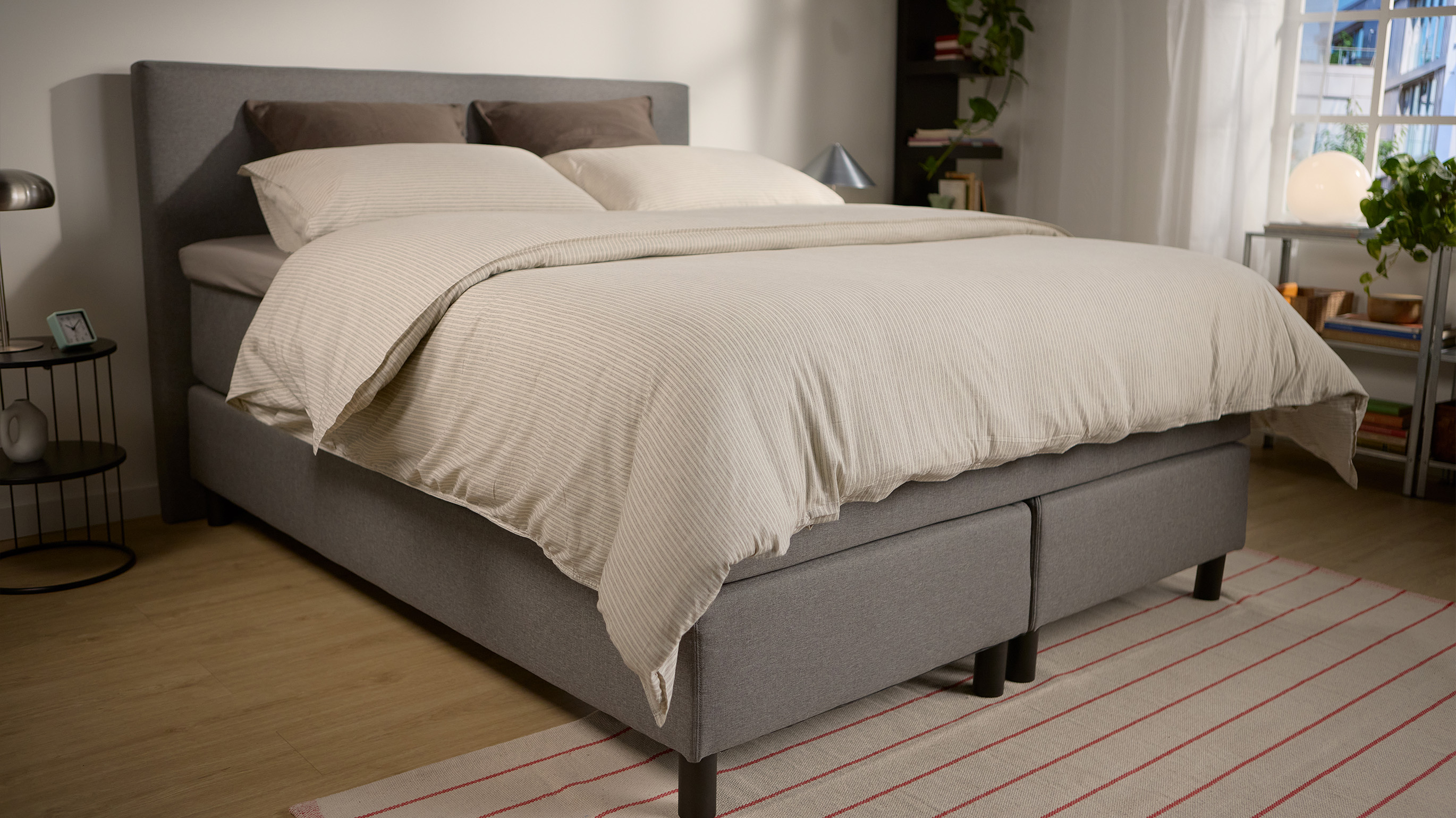 Emma Boxspring Bed in grey with upholstered headboard and dark legs, styled with cream striped bedding in a bright bedroom.
