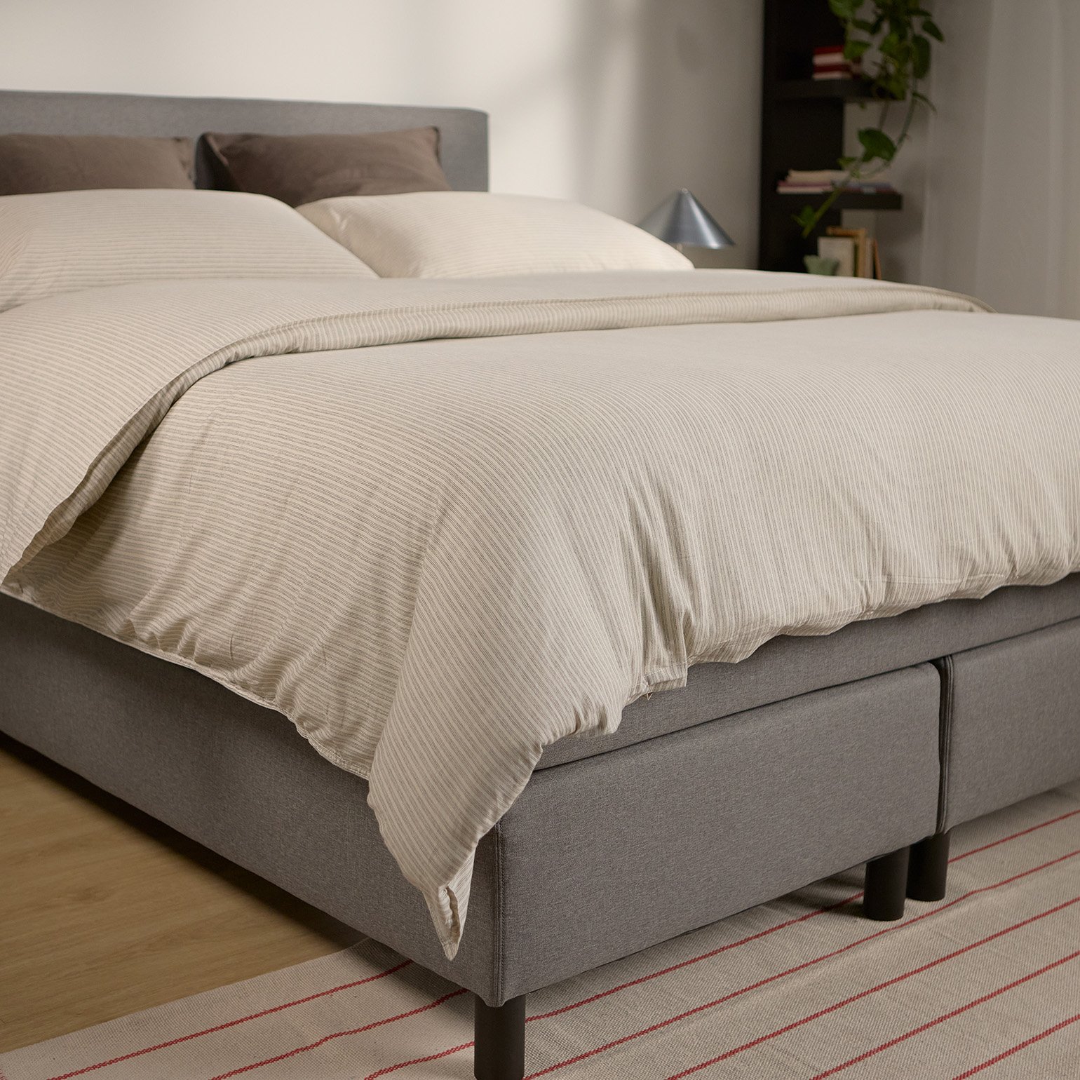 Emma Boxspring Bed in grey with upholstered headboard and dark legs, styled with cream striped bedding in a bright bedroom.
