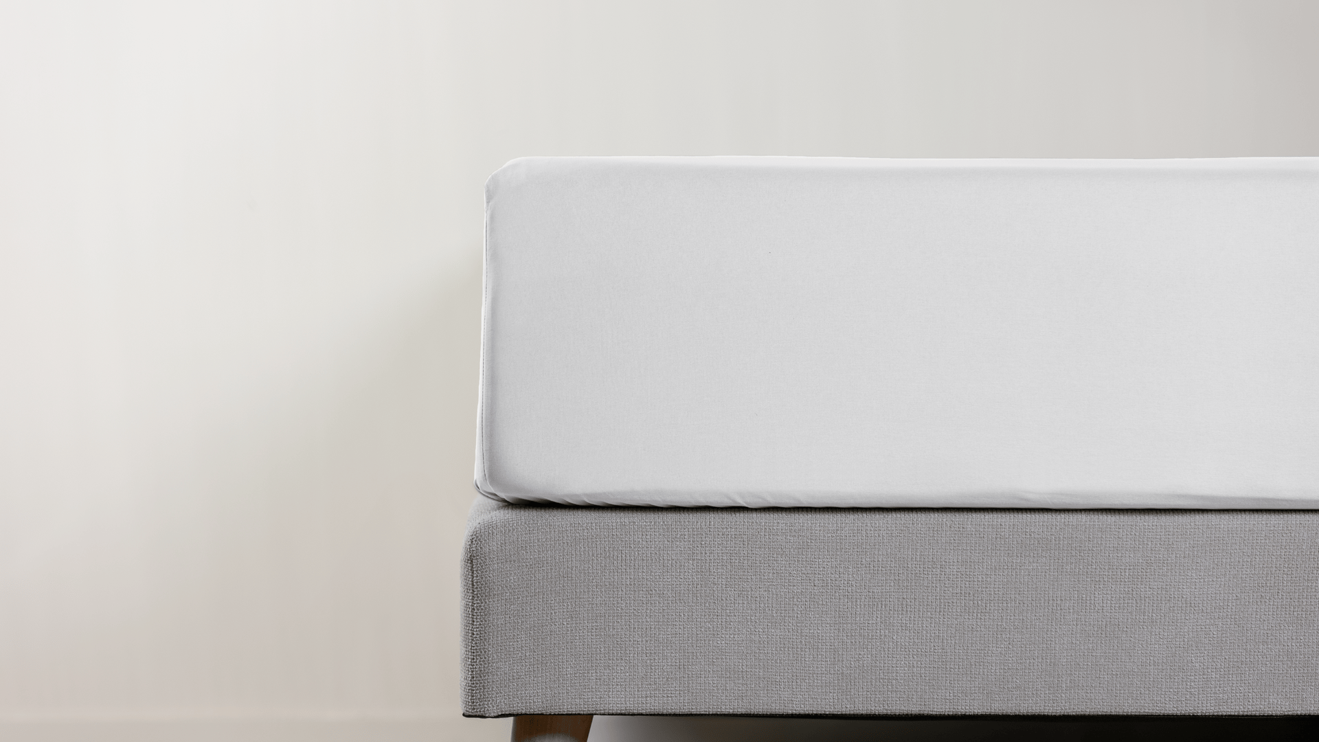 Slaapkamer met een opgemaakt bed met lichtgrijs lakens, een houten nachtkastje met lamp en een plant ernaast.