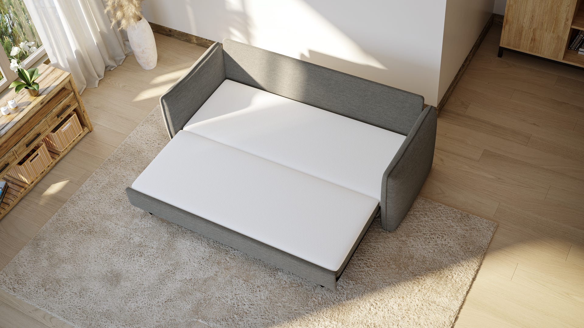 Canapé convertible gris déplié en surface de couchage, posé sur un tapis clair dans un salon minimaliste. Lumière naturelle entrant par une fenêtre avec rideaux blancs à gauche. Table basse en bois style palette avec plante en pot. Ambiance cosy, épurée et fonctionnelle.