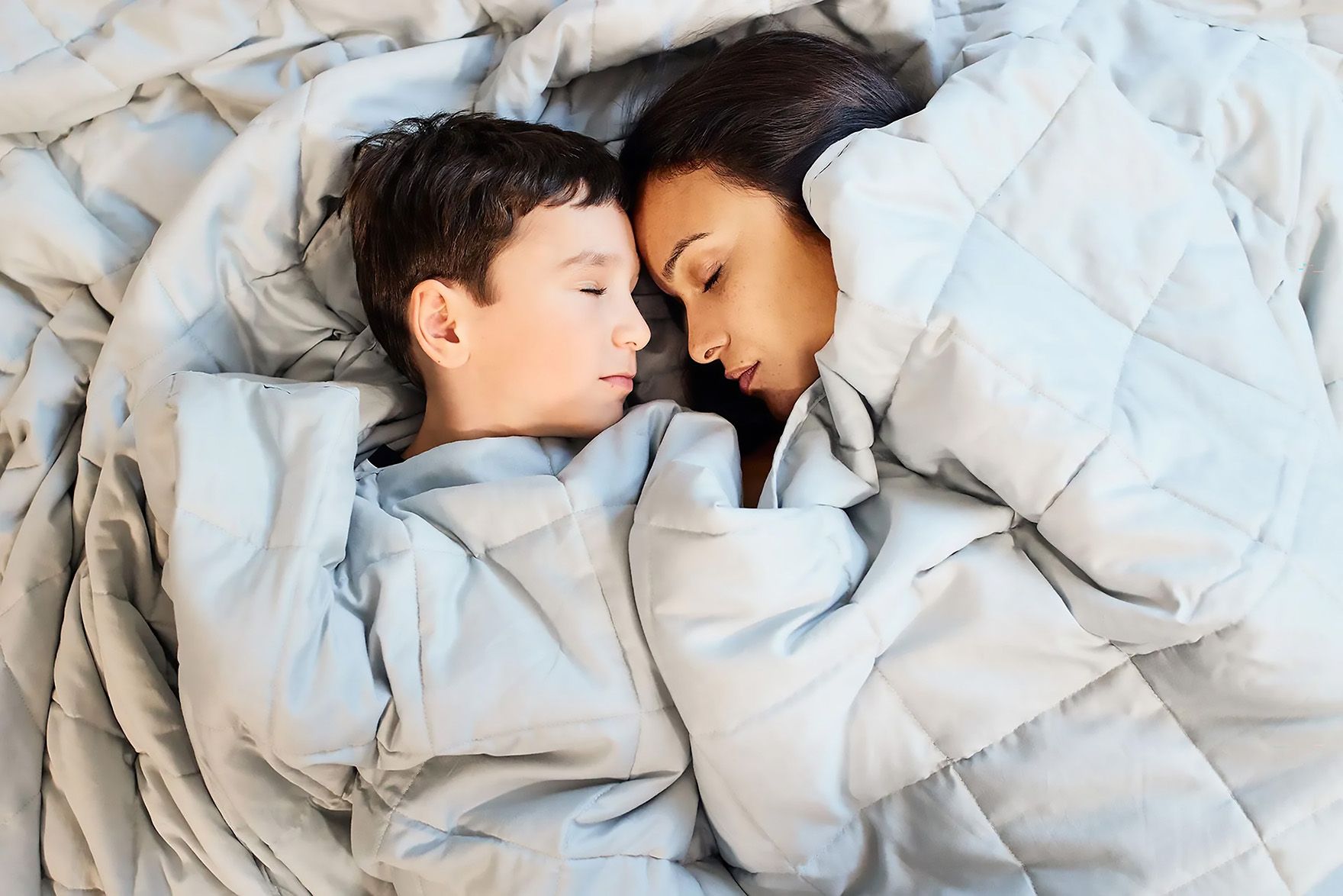 Mutter und Sohn schlafen friedlich nebeneinander unter einer hellgrauen Bettdecke.