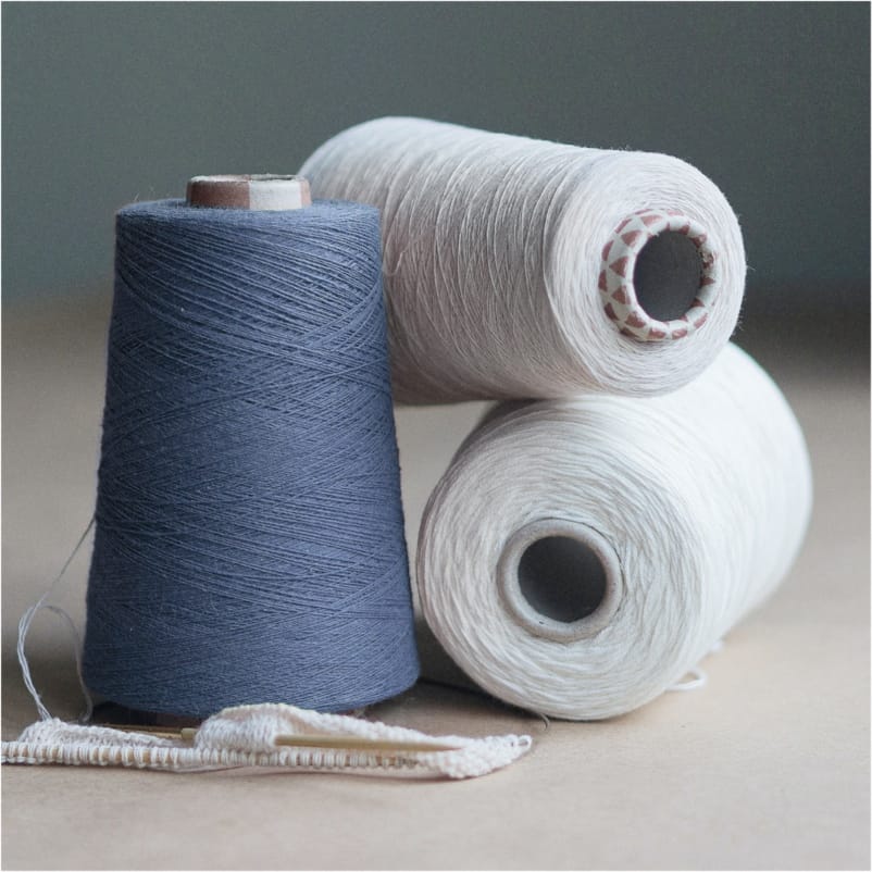 Drie klossen garen—één donkerblauw rechtop en twee witte liggend—naast wit textiel met naald, op oppervlak voor naaien of textielbewerking.