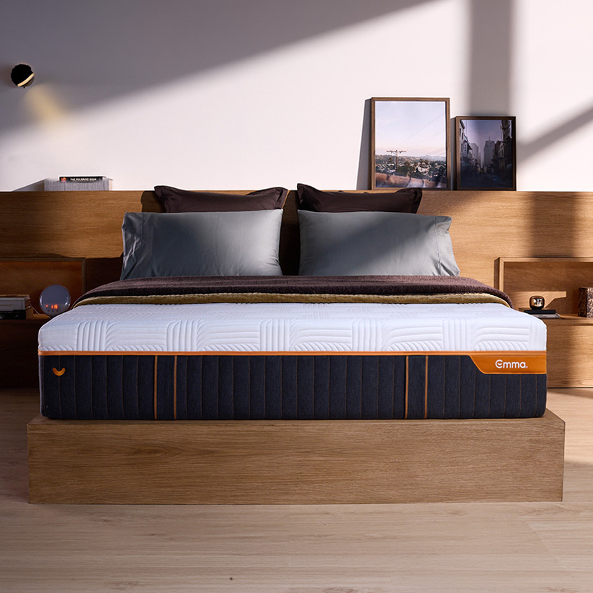 Front view of mattress in a room, showcasing it's height, dark base and white cover, with the orange Emma logo and zipper. 