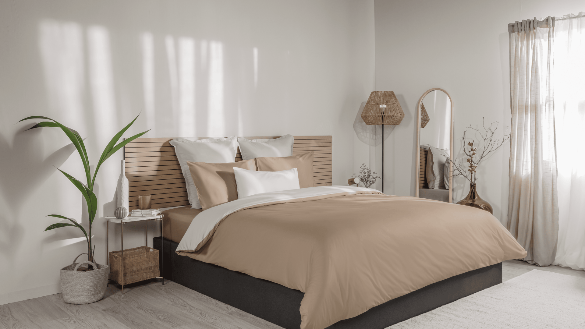 Emma Beddengoed Set Percal in kleurvariant Crème & Beige, bestaande uit een dekbedovertrek en twee kussenslopen, strak opgemaakt op een bed met houten lattenhoofdbord; links een houten nachtkastje met vaas, kaars en boeken, daarnaast een groene kamerplant; rechts een grote spiegel en een rieten vloerlamp; lichte slaapkamer met houten vloer en transparante gordijnen die zacht daglicht doorlaten.