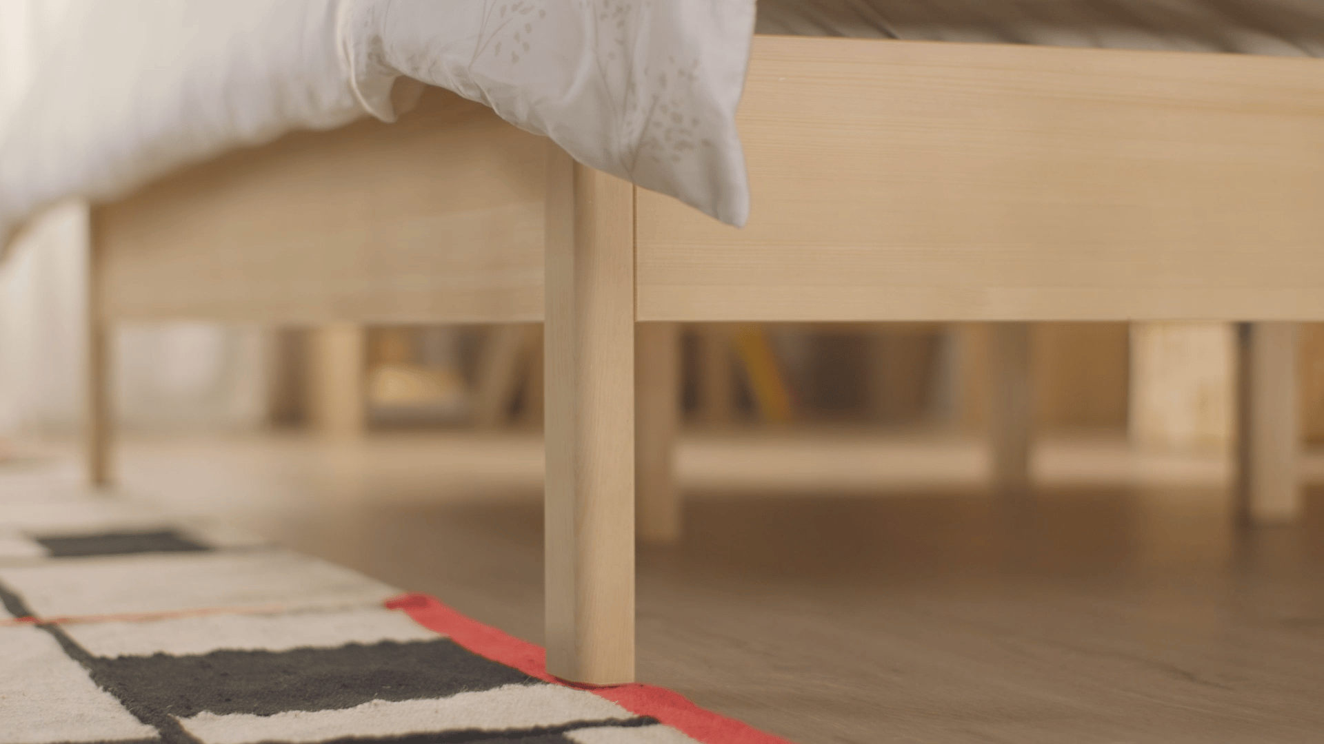 Licht houten Emma bedframe met witte dekbed en subtiele bloemenprint, gefotografeerd vanuit laag perspectief met zicht op de poten en een zwart-wit-rood vloerkleed op lichte houten vloer.