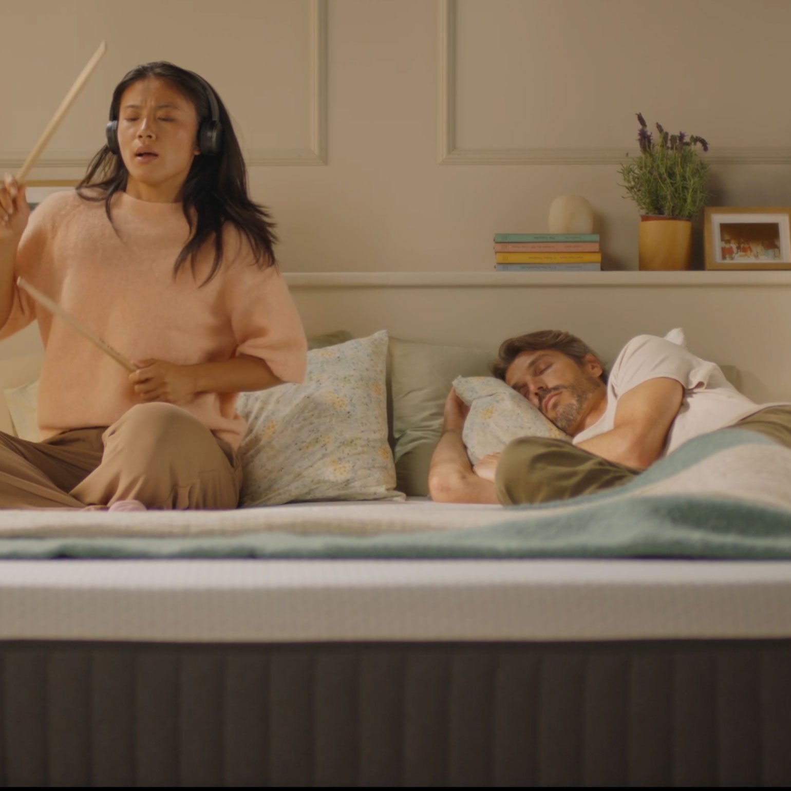 Person air-drumming with headphones on an Emma Original mattress while their partner sleeps undisturbed beside them.