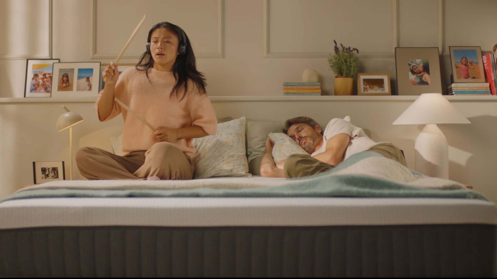 Person air-drumming with headphones on an Emma Original mattress while their partner sleeps undisturbed beside them.