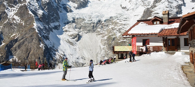 Personen auf Ski und Snowboard vor Berghütte im Aostatal.